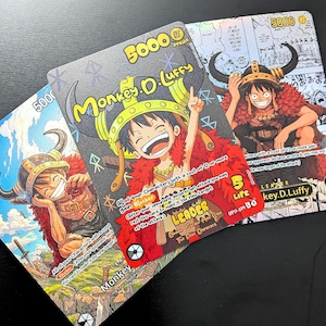 Pode incluir: Três cartas colecionáveis de One Piece com Monkey D. Luffy como líder. As cartas exibem ilustrações coloridas em estilo mangá do personagem usando um capacete com chifres e capa vermelha, com texto incluindo 5000 de poder e 5 de vida.