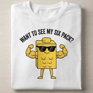 May include: A white t-shirt featuring a graphic of a yellow, block-shaped character wearing black sunglasses. The character has muscular arms and a six-pack abdominal design. Above the character, the text reads: WANT TO SEE MY SIX PACK?