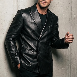 May include: A black double-breasted leather blazer featuring a notched lapel, long sleeves with buttoned cuffs, and two front flap pockets. The jacket is worn over a plain black t-shirt, showcasing a smooth, polished finish and structured tailoring.