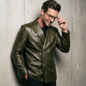 May include: A double-breasted olive green leather blazer featuring a notched lapel, long sleeves with buttoned cuffs, and a structured fit. The jacket is worn over a dark shirt and paired with thin-rimmed glasses with brown frames.