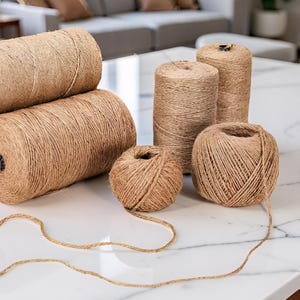 Puede incluir: Varios carretes y bolas de hilo marrón natural están dispuestos sobre una superficie de mármol blanco. El hilo es de varios tamaños, incluyendo rollos grandes y bolas más pequeñas, con un trozo de hilo que se extiende desde una de las bolas.