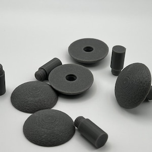 May include: A collection of gray, 3D-printed hardware components. The assortment includes cylindrical posts with threaded ends, flat circular pieces with central holes, and rounded, knob-like elements. The pieces are arranged on a white surface.
