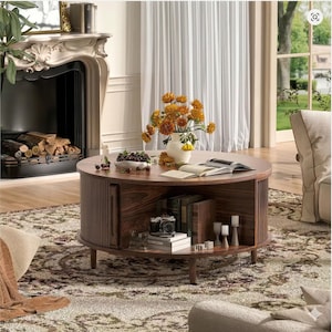 May include: A round, dark wood coffee table with a slatted side panel and a lower shelf. The table holds books, a camera, and candles. A vase of orange flowers sits on top, along with open books and a bowl of fruit. The table is on a patterned rug.