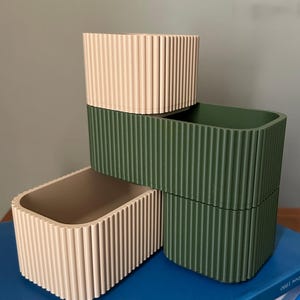 Puede incluir: Tres contenedores rectangulares de almacenamiento en beige y verde oliva están apilados sobre una superficie azul. Cada contenedor tiene un exterior acanalado y esquinas redondeadas. Los contenedores están vacíos y parecen ser de plástico.