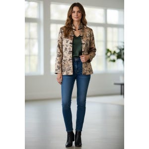 May include: A woman wearing a beige floral print jacket with black frog closures, a dark green top, and blue jeans. The jacket has a mandarin collar and long sleeves. She is also wearing black ankle boots.
