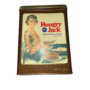 May include: Vintage Hungry Jack Buttermilk Complete Pancake Mix tin. The tin features a smiling woman in a vintage dress, with the words "Hungry Jack" in red and "Pillsbury" in blue. The tin is brown with a gold trim.