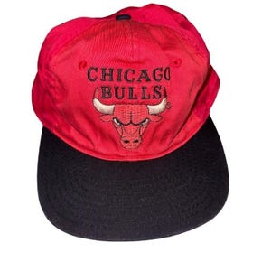 May include: A red and black Chicago Bulls baseball cap. The cap displays the team's logo and the words "CHICAGO BULLS" embroidered in black. It has a curved peak and an adjustable strap.