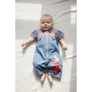 May include: A baby in a blue denim jumpsuit with a red and white checked collar and sleeves. The jumpsuit has a white zip and a cartoon cowboy graphic. The text "Howdy Partner" is printed on the jumpsuit.