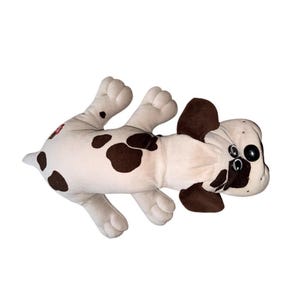 May include: A plush toy dog in a lying down position. The dog is primarily white with brown spots and accents on the ears and around the eyes. It has large, black button eyes and a small red tag.