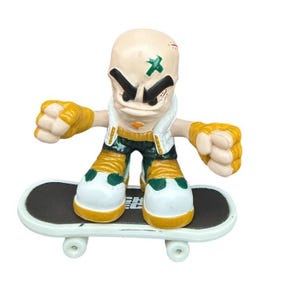 May include: A small, stylized figurine of a bald character with an angry expression, wearing a white vest, green and gold accents, and yellow gloves. The figure is riding a white skateboard with a black deck. The character has a green cross on its head.