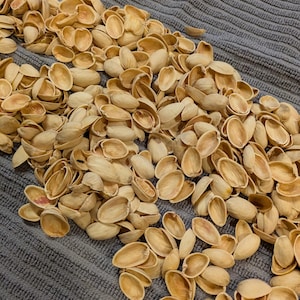 May include: A pile of empty pistachio shells, beige in color, scattered on a textured gray surface. The shells are open, revealing the inner surface. The image is a close-up, showing the details of the shells and the fabric.