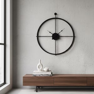 Op de afbeelding: Een grote, zwarte, minimalistische wandklok met een cirkelvormig frame en kruisvormige steunen. De klok heeft zwarte wijzers en een centrale naaf. De klok is gemonteerd op een getextureerde, lichtgrijze muur. Een houten kast met decoratieve items staat eronder.