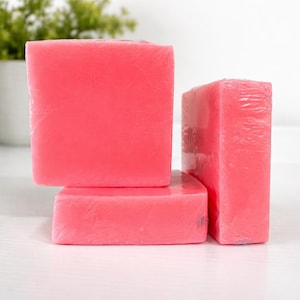 Könnte beinhalten: Drei rechteckige, rosa Seifenstücke sind gestapelt. Die Seife hat eine kräftige, rosa Farbe und scheint in klarem Kunststoff verpackt zu sein. Die Riegel sind unterschiedlich groß, mit einem größeren Riegel oben.