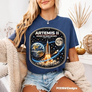 May include: Navy blue t-shirt featuring a graphic of the Artemis II mission, with a rocket launching from Earth towards the moon. The design includes the text "ARTEMIS II AROUND THE MOON AND BACK APRIL 1, 2026".