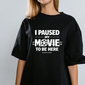 May include: Black t-shirt with white text: "I PAUSED MY MOVIE TO BE HERE" and a film reel graphic. The phrase "priorities matter." is below the text.