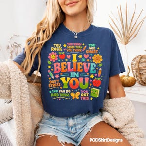 Könnte beinhalten: Marineblaues T-Shirt mit dem Text "I Believe in You" in bunten Buchstaben. Das Shirt zeigt verschiedene Grafiken, darunter Blumen, Äpfel, Bleistifte und die Sätze "You Rock", "You are Smart" und "You Got This".