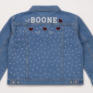 May include: A light blue denim jacket featuring the name "BOONE" in silver lettering on the upper back. The jacket is adorned with sparkling rhinestones and red heart-shaped patches. A classic, stylish outerwear piece.