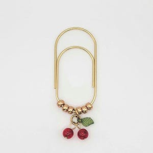 May include: A gold-colored paper clip with a cherry charm. The paper clip has a rounded rectangular shape with a series of small, gold-colored beads at the bottom. A charm of two red cherries and a green leaf hangs from the beads.