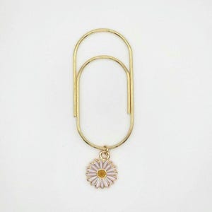 May include: Gold-colored paper clip with a daisy charm. The daisy charm has a pink and white petal design with a yellow center. The paper clip is made of a thin, curved metal.