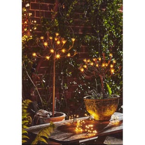 May include: Outdoor decorative lights shaped like starbursts, with warm yellow lights, are displayed in pots and on a wooden table. The lights are set against a brick wall and greenery, creating a cozy ambiance.