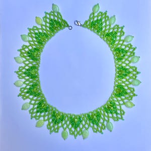 Op de afbeelding: Handgemaakte kralenketting in groentinten. De ketting heeft een delicaat netachtig ontwerp met kleine groene kralen en grotere, ovaalvormige lichtgroene kralen. Het heeft een zilverkleurige sluiting.