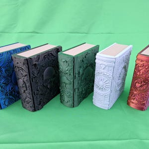 Könnte beinhalten: Fünf dekorative, buchförmige Boxen in einer Reihe, jede mit einer anderen Farbe und einem anderen Design. Die Farben sind Blau, Schwarz, Grün, Weiß und Rot. Die Boxen haben aufwendige Prägemuster und eine hellfarbige Oberkante.