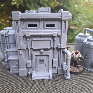 Può includere: Un modello di edificio industriale grigio stampato in 3D con una struttura centrale dotata di porta, tubi e prese d'aria, accompagnato da due serbatoi cilindrici e una piccola miniatura dipinta su una base circolare sul lato destro.