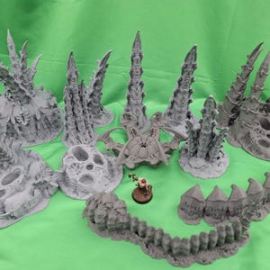 Peut inclure: Ensemble de pièces de terrain grises imprimées en 3D pour les jeux de table, comprenant des flèches, des structures en forme de crâne et un mur segmenté. Une figurine miniature se trouve près du terrain, sur fond vert.