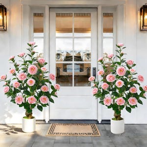 May include: Two artificial camellia trees with pink blossoms in white pots flank a white door. The trees have green leaves and brown trunks. The pots sit on a gray concrete surface, with a decorative doormat in front of the door.