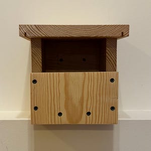 May include: A wooden birdhouse with a rectangular body and a small roof. The birdhouse is made of light-colored wood and has a natural wood grain. The birdhouse is mounted on a white wall.