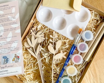 May include: A craft kit with wooden flower cutouts, paint, brushes, and a palette. The kit includes a variety of paint colors, wooden tools, and a small envelope. The items are arranged in a brown box with straw.