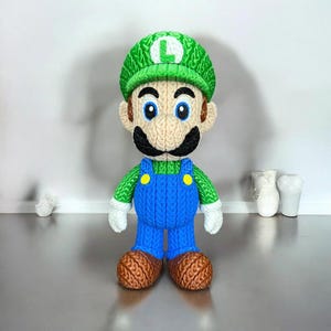 May include: A knitted Luigi doll, with a green hat bearing a white "L", a black moustache, and blue dungarees. The doll has brown shoes and is made with textured yarn.