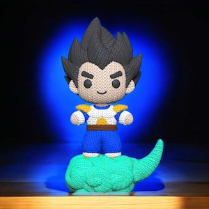 May include: A knitted figure of a character with spiky black hair, wearing a blue and white outfit with yellow accents. The figure stands on a green cloud-like base. The background is dark blue.