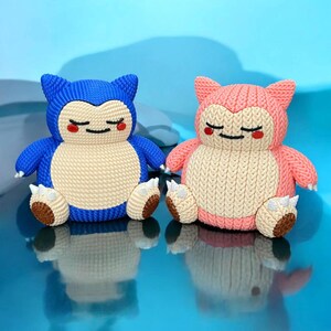 Könnte beinhalten: Zwei Plüschfiguren, eine blaue und eine rosa, die einer Zeichentrickfigur ähneln. Sie haben eine Strickstruktur, cremefarbene Bäuche, braune Pfoten und geschlossene Augen. Die Figuren sitzen vor einem hellblauen Hintergrund.