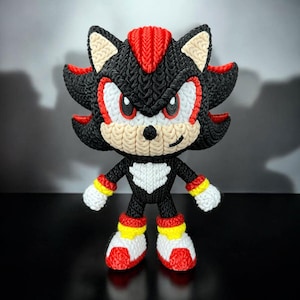 May include: A handmade, knitted Shadow the Hedgehog figure. The figure is primarily black with red, white, and yellow accents. The hedgehog has a detailed knitted texture, with red spikes, white chest, and yellow and red details on the arms and feet.