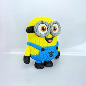 May include: A yellow Minion figure with blue overalls, black shoes, and gray goggles. The figure has a knitted texture and a smiling expression with a pink tongue. The Minion is a popular character from the Despicable Me franchise.