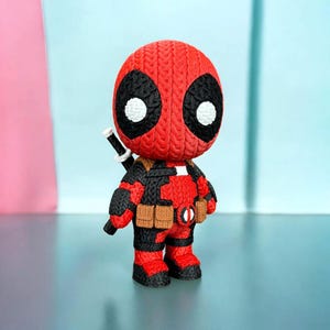 May include: A red and black Deadpool figurine with a knitted texture. The figure has large black eye shapes with white circles, a utility belt, and a sword on its back. The figure is standing on a reflective surface.