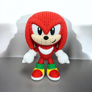 May include: A knitted red Knuckles the Echidna figurine with white accents on the face, hands, and chest. The figure has green and yellow shoes. The character is from the Sonic the Hedgehog video game franchise.