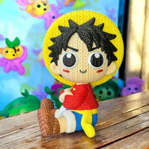 May include: A knitted figurine of a cartoon character with black spiky hair, a yellow hat, and a red shirt. The figurine is sitting on a wooden surface. The background is blurred, with other colourful figurines visible.