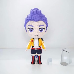 May include: A handmade crochet doll with a purple hairstyle, large eyes, and a smiling face. The doll wears a yellow and white jacket, dark blue shorts, and red and black boots. A small, white, rectangular object and a silver stand are in the background.