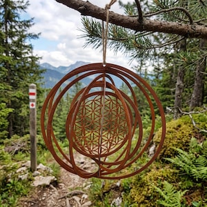 May include: A rusty brown metal ornament with a geometric design, hanging from a tree branch. The ornament features concentric circles and a central pattern of overlapping circles. The background shows a mountain landscape with trees and a hiking trail.
