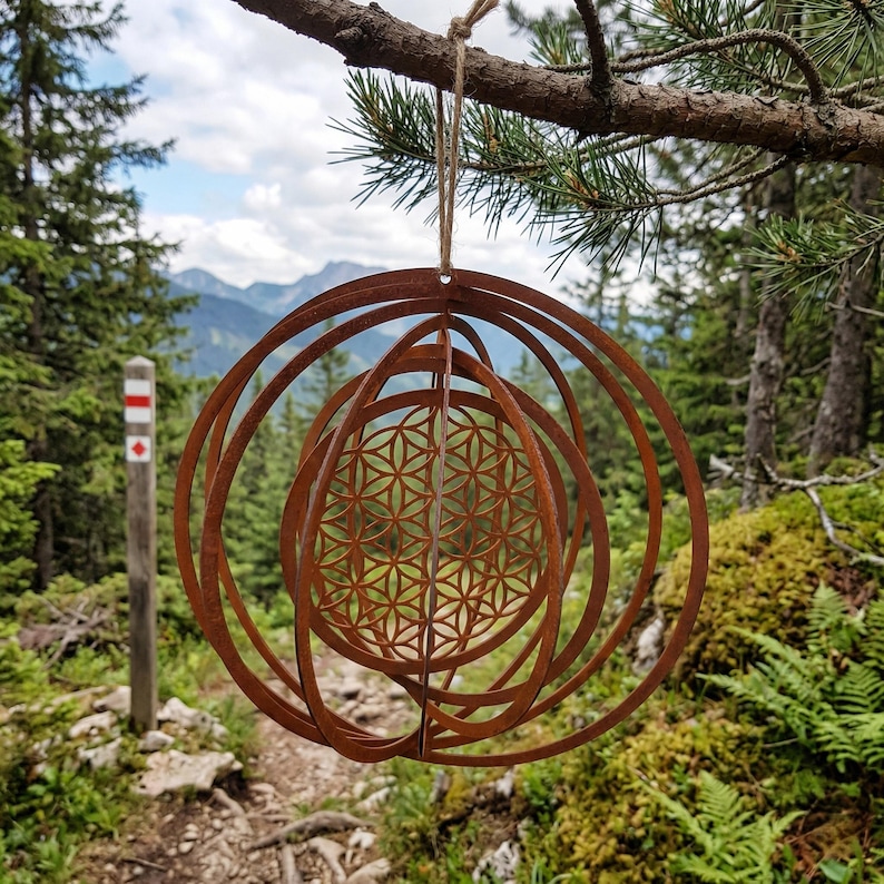 May include: A rusty brown metal ornament with a geometric design, hanging from a tree branch. The ornament features concentric circles and a central pattern of overlapping circles. The background shows a mountain landscape with trees and a hiking trail.