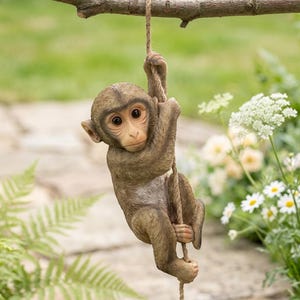 May include: A brown monkey figurine hanging from a rope, attached to a tree branch. The monkey has large, expressive eyes and is sculpted with realistic details. The background features green foliage and white flowers, creating a natural setting.