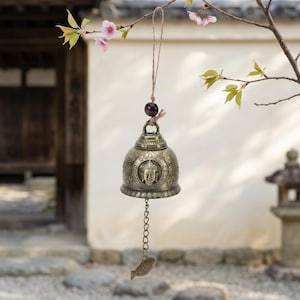 Peut inclure: Une cloche de carillon éolien de couleur bronze avec un motif de visage de Bouddha, suspendue à une branche avec des fleurs roses. La cloche a une chaîne avec un charme en forme de poisson. L'arrière-plan montre un bâtiment traditionnel.
