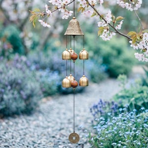 Peut inclure: Un carillon en bronze avec un sommet en forme de cloche et plusieurs petites cloches suspendues en dessous. Des perles en bois sont intercalées entre les cloches. Une pièce décorative est suspendue en bas. Le carillon est suspendu à une branche d'arbre avec des fleurs blanches.