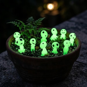 May include: A terracotta plant pot filled with dark soil, a small green fern, and eleven small, glowing, light green figurines. Each figurine features a smooth, rounded head with two black circular eyes and a simple, minimalist body shape.