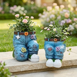 Op de afbeelding: Twee plantenbakken in de vorm van een jeansbroek, elk gevuld met bloemen. De plantenbakken hebben een versleten uitstraling met patches en een bruine riem. Eén plantenbak heeft roze rozen en madeliefjes, de andere lavendel en madeliefjes. Ze staan op een stenen oppervlak.