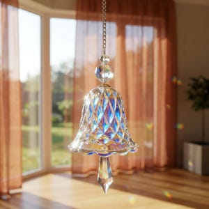 Puede incluir: Una campana de cristal transparente y facetado colgando de una cadena. La campana tiene un patrón de diamantes y una base puntiaguda. El cristal refleja la luz, creando un efecto arcoíris. La campana está suspendida frente a una ventana.