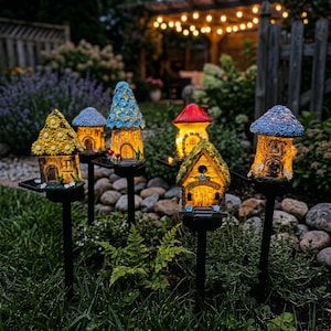Puede incluir: Una colección de estacas de jardín con forma de casa de hadas iluminadas. Cada casa presenta detalles únicos, incluyendo diseños de flores, setas y tejados texturizados. Las estacas están colocadas en un jardín con un fondo borroso de vegetación y luces de cadena.