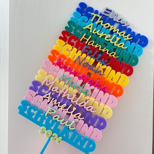 Könnte beinhalten: Ein mehrfarbiger Acryl-Tortentopper mit dem gestapelten Schriftzug SCHULKIND in verschiedenen Farben und darauf liegenden Namen in Schreibschrift, darunter Elias, Thomas, Aurelia, Hanna, Felix, Noah, Franziska, Merle, Mathilda, Amelia, Paul und 2026.